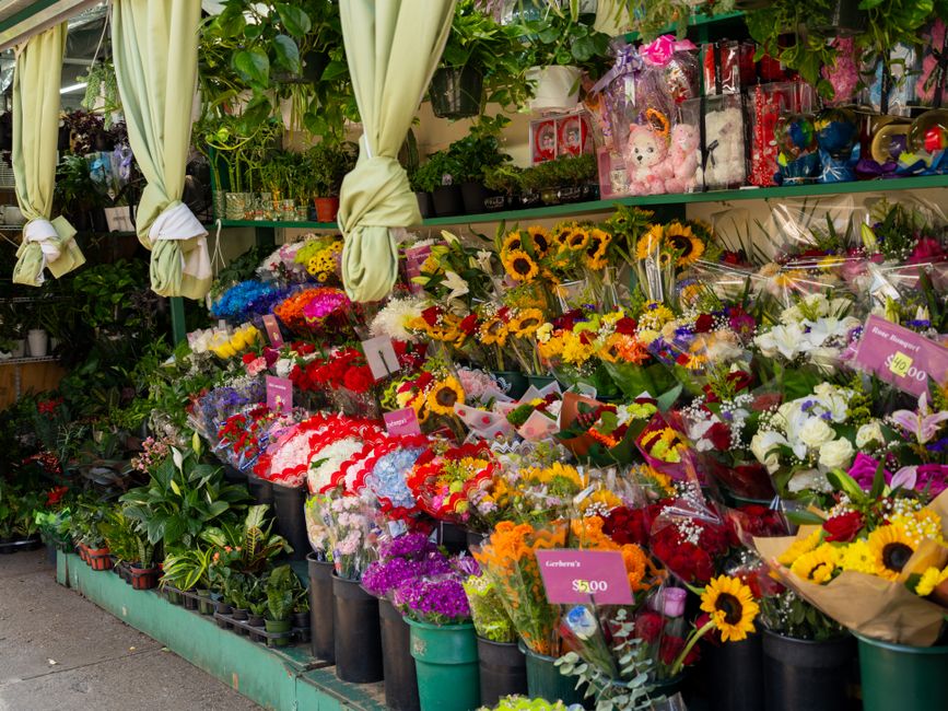 A vibrant flower display outside a florist shop showcasing a variety of fresh floral arrangements, including sunflowers, roses, lilies, and mixed bouquets wrapped in clear cellophane and decorative pa