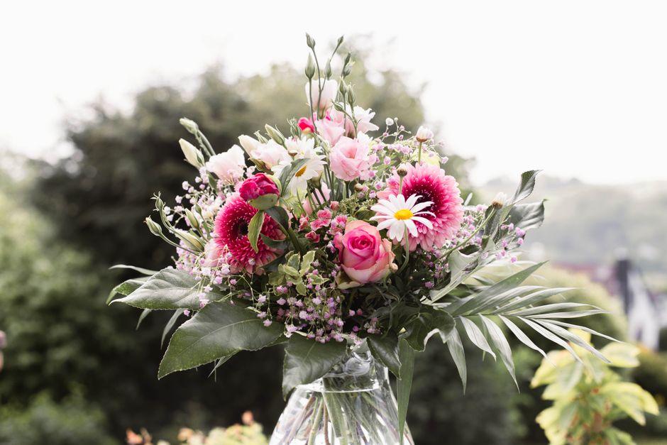 A vibrant floral bouquet arranged in a clear glass vase, featuring pink roses, white daisies with yellow centers, pink gerbera daisies, and sprigs of delicate pink fillers, accented with lush green fo