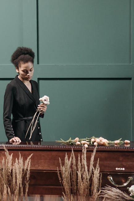 A young woman with dark curly hair styled in an updo and light skin, standing upright next to a polished wooden casket in a dimly lit room. She is wearing a black blazer with metallic button details a
