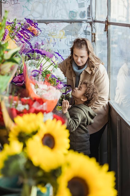 A woman with wavy light brown hair, wearing a beige coat over a blue top, is helping a young boy with curly hair select flowers inside a greenhouse. The boy, dressed in a dark green hoodie, is reachin